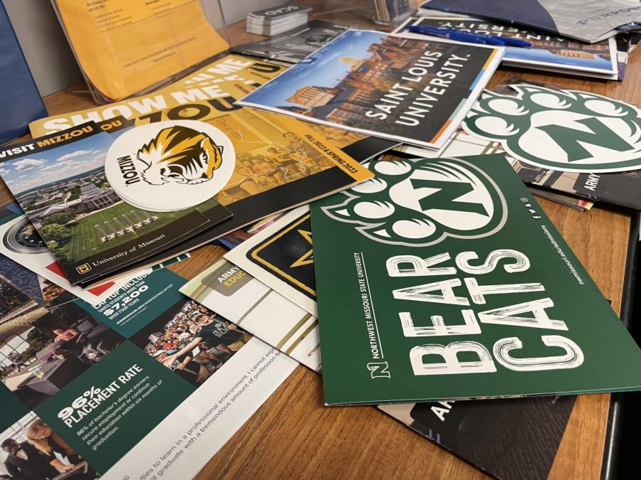Various college brochures sit scattered in a pile across a table. 