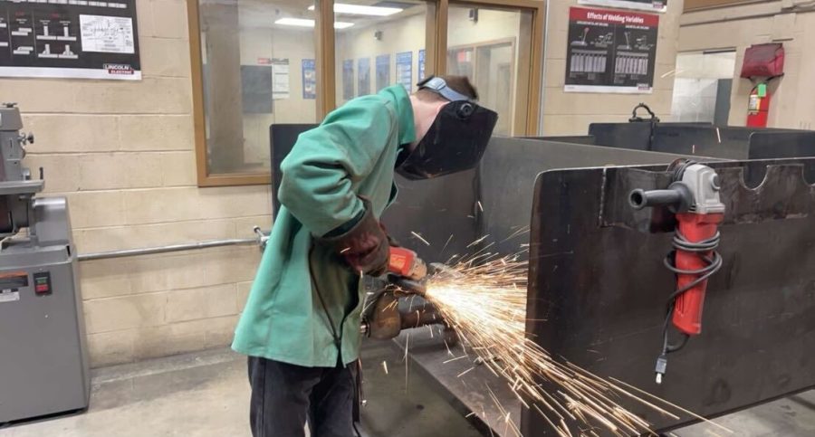 A welding student grinds the debris off of a weld while the metal is secured by a table vice 