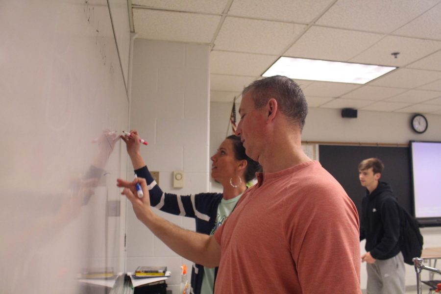 Teachers Lindsey and Larry Scheller write on whiteboards. The Schellers are married and have been working together for over 17 years.  Lindsey teaches classes such as The Novel, as well as English I and II. Meanwhile, Larry teaches biology, anatomy and physiology