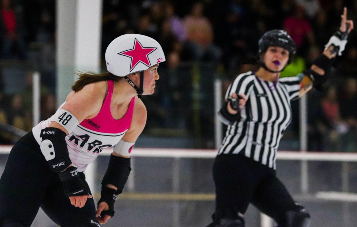 Number 48 on the white team looks ahead as she skates around the track during a scrimmage on Feb. 17. A ref holds up their arm in the background, signaling the skater as the lead jammer. The lead jammer in a given jam is the first player with a star on their helmet to break through the large group of blockers, also known as the pack. Being signified as lead jammer gives that player the ability to end the jam at any time by tapping their hips.

I was a skater for seven years, outside pack referee Kim Mason said. I started playing in 2012 and I had heard about it for years before that. My job tonight was to watch the skaters from the outside of the track and watch for penalties.
