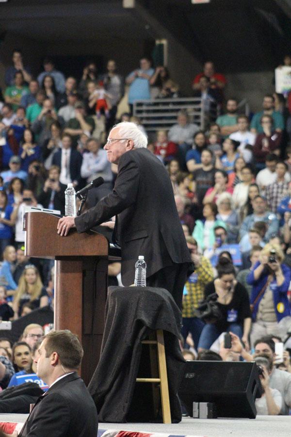 Senator Sanders stands at his podium while he speaks. Sanders stood on a stage overlooking the crowd and surrounded by police officers, bodyguards, and secret service agents. 
