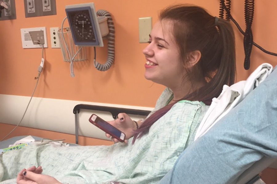 Junior Kailey Cripps sits in the hospital, smiling, while playing on her phone.