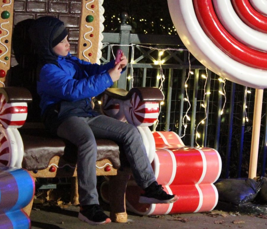 A little boy bundled up in his winter attire takes a selfie on his mother’s phone as he sits on the gingerbread inspired chair. This is one of the many locations where families can take group photos or rest in front of the lake. 