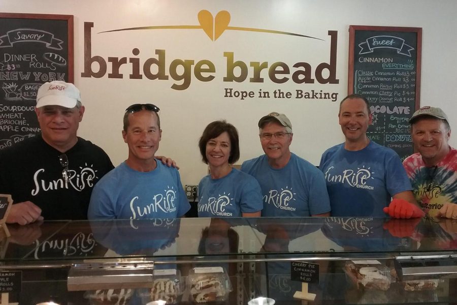 The owners of Bridge Bread stand in a line behind the counter as they pose for a picture. Bridge Bread is an organization that feeds the homeless and gives them jobs. They bake bread, bagels, cake and brownies daily.