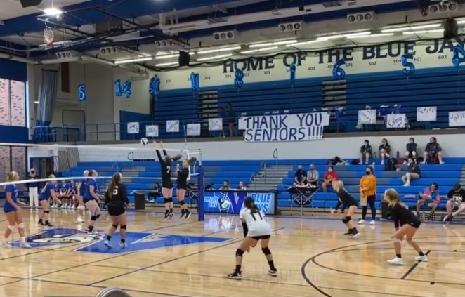 Player number seven, Sara Ausbury, jumps up tall with her teammate, Megan Steutermann as they  raise both arms up, careful not to touch the net, to attempt to block the opponents hit. 