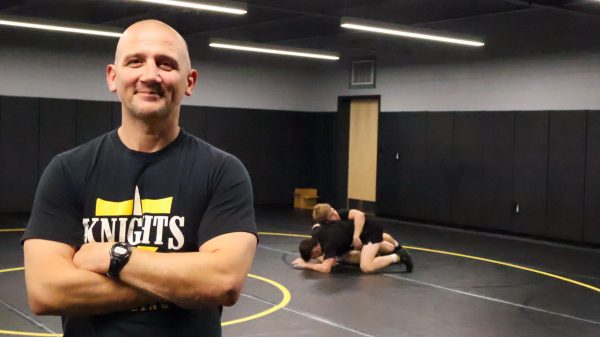 Wrestling head coach, Chris Brown, poses for a photo while two wrestlers compete in the background.